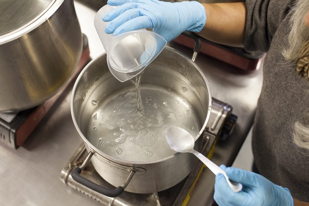 Pouring clear liquid from a plastic beaker into a metal pot on a burner.
