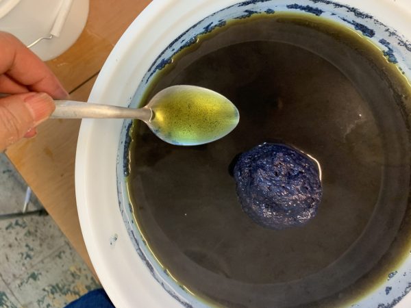 A spoonful of bright yellow liquid above an indigo vat with an opaque copper sheen on top.