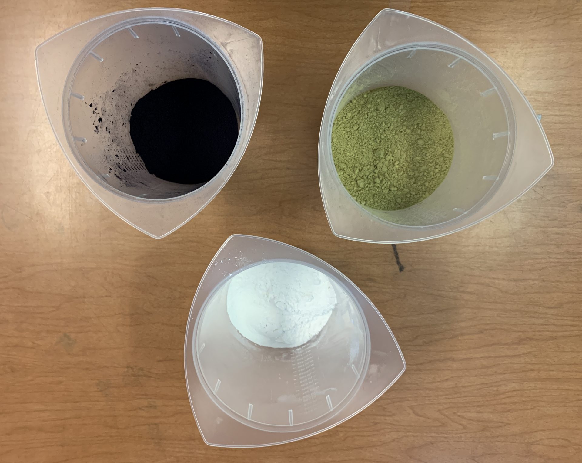 Three plastic beakers with powders in them. One has a dark blue powder, one has a yellow-green powder, and one has a white powder.