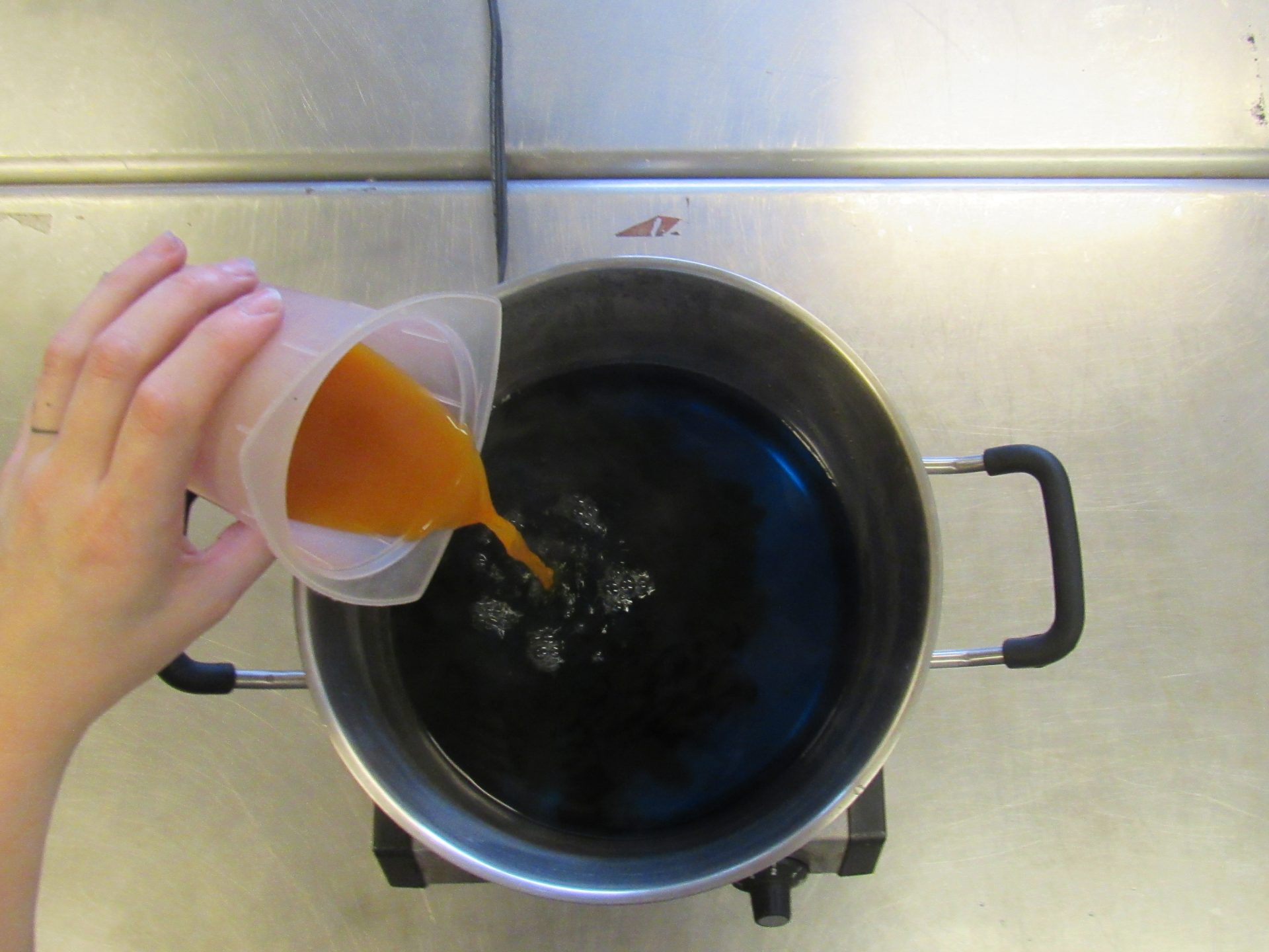 a hand pouring an orange liquid from a plastic beaker into a dyepot with blue liquid in it. You can see the two colors mixing in the dyepot.