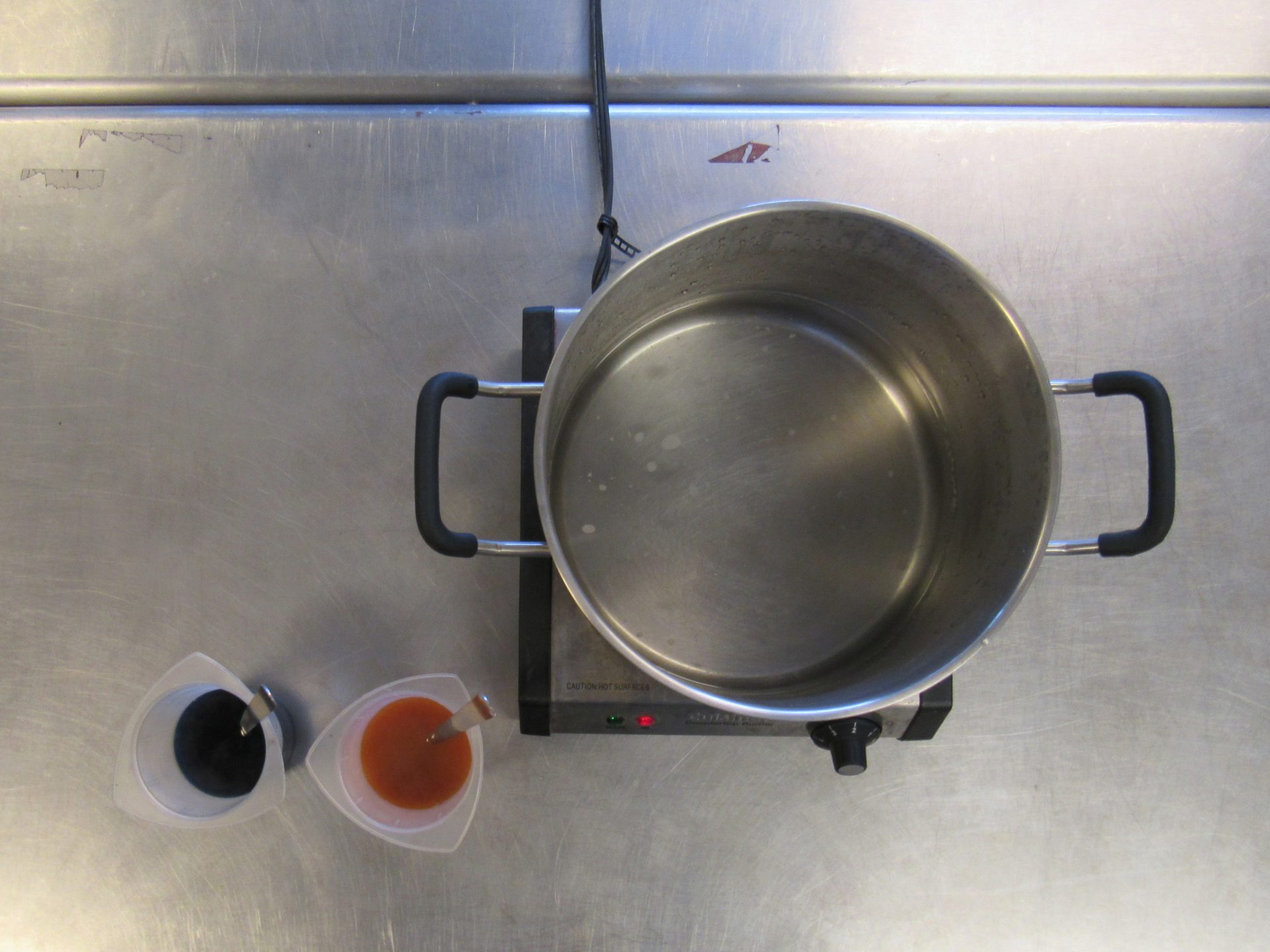 Two beakers with colored liquid dyes next to a pot with water on a burner.