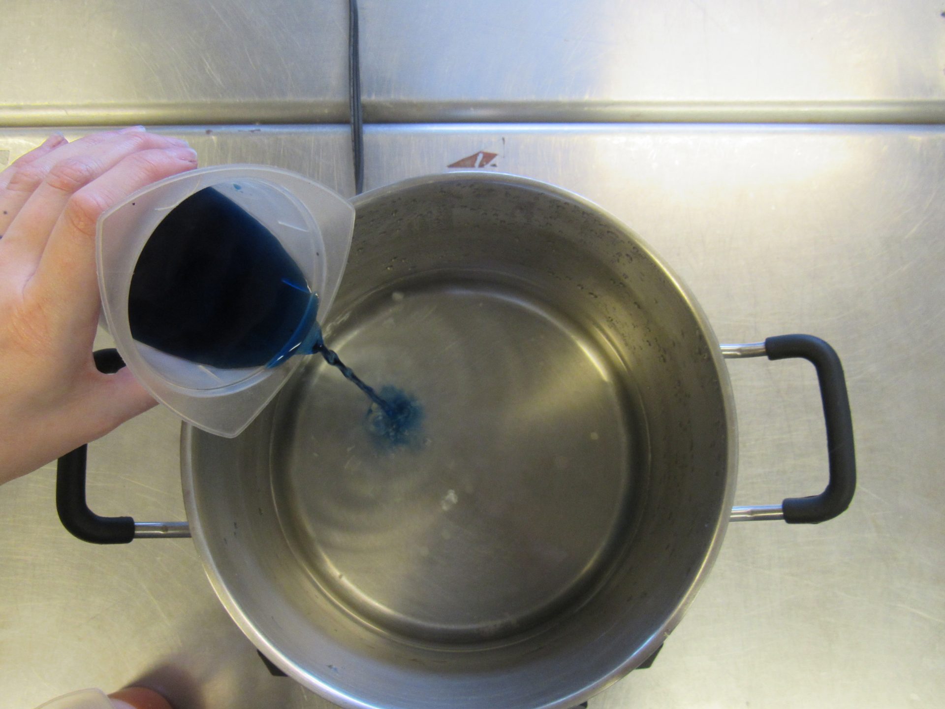 a hand pouring a beaker of bright blue liquid into a pot