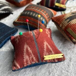 Handwoven square petite pincushion in an orange colored pattern with pins and a small bronze plawue that says Threads of Life