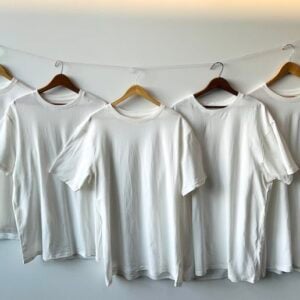 Five mordanted organic t-shirts are hanging on wooden hangers facing front. They are all white against a white background.