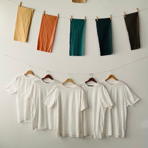 Five white organic mordanted t-shirts hang from wooden hangers facing front with five colored cotton banners hanging above them